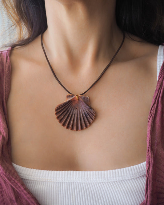 Model wearing a necklace with a big scallop shell on a brown suede cord. The shell is a great scallop shell from Portugal and is about 6 cm. The color of the shell is bordeaux brown and it has vertical ridges. 