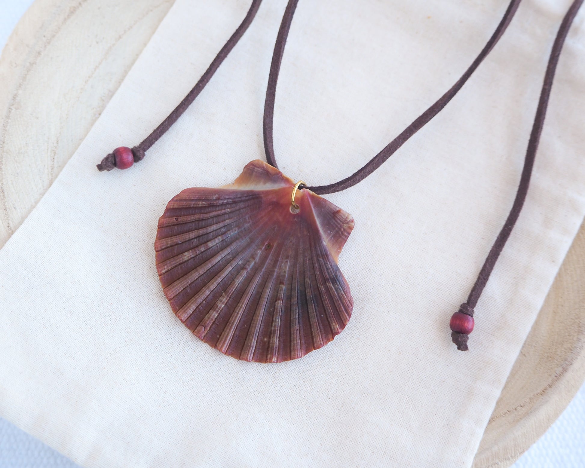 Side view of a necklace with a big scallop shell on a brown suede cord on light cotton displayed. The shell is a great scallop shell from Portugal and is about 6 cm. The color of the shell is bordeaux brown and it has vertical ridges. 