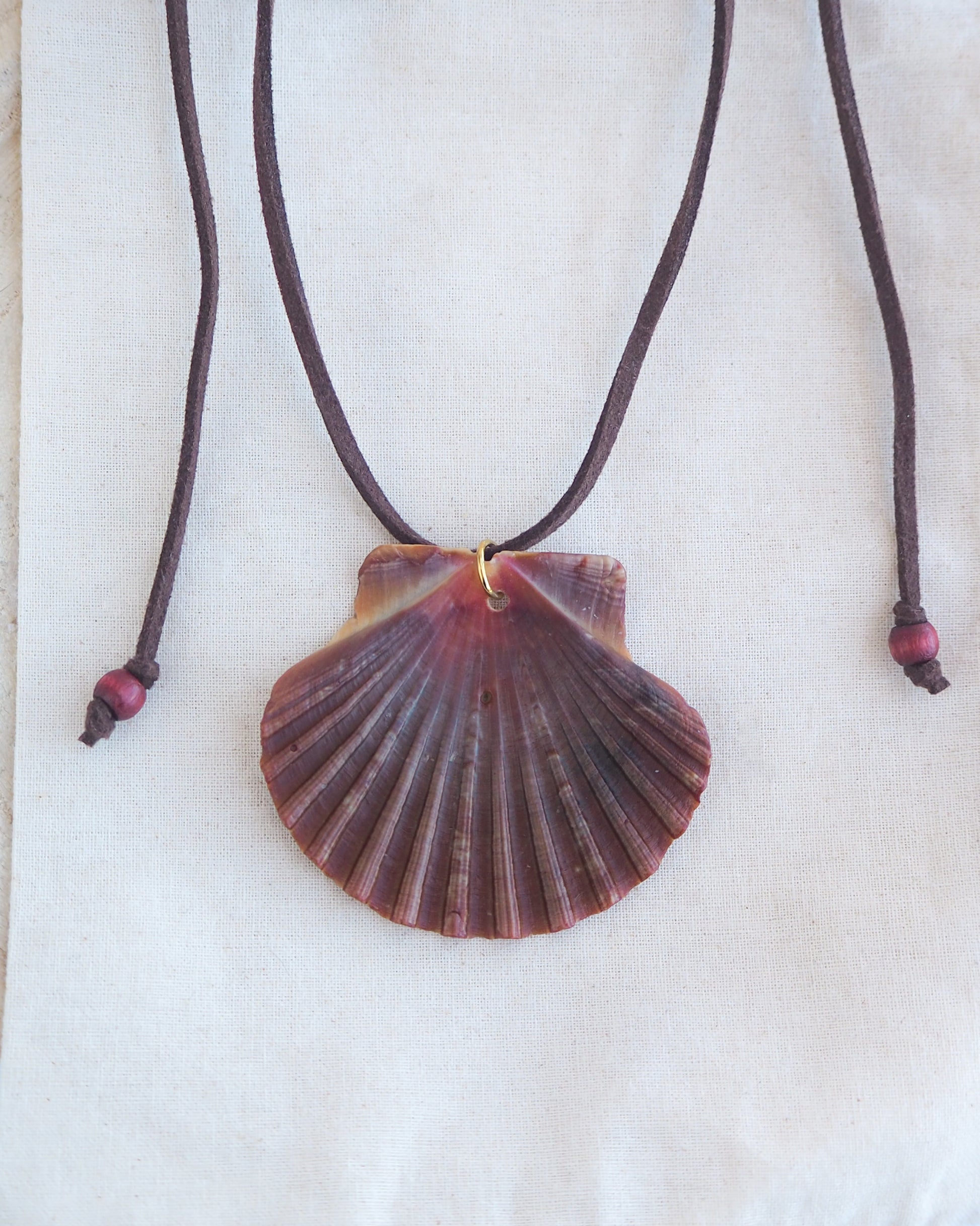 Front view of a necklace with a big scallop shell on a brown suede cord on light cotton displayed. The shell is a great scallop shell from Portugal and is about 6 cm. The color of the shell is bordeaux brown and it has vertical ridges. 