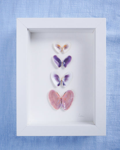 Front view of Artwork with butterfly-shaped seashells in various colors on a light gray background. Made with handpicked shells from Portugal. Coquina shells and Gari Virgata Shells. Set in a white frame box.