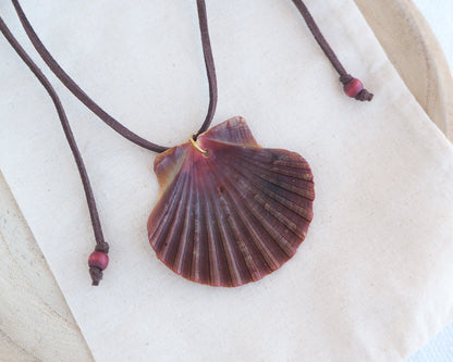 a necklace with a big scallop shell on a brown suede cord on light cotton displayed. The shell is a great scallop shell from Portugal and is about 6 cm. The color of the shell is bordeaux brown and it has vertical ridges. 