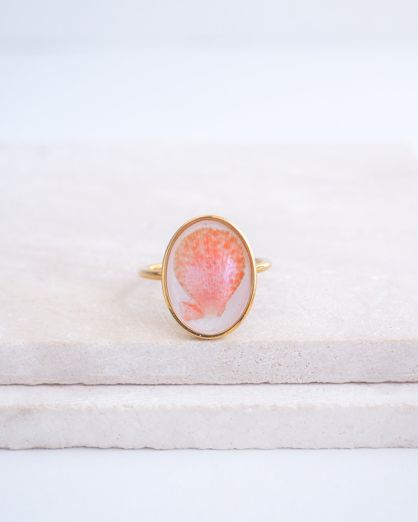 Ring with a pink and gold design on a white fabric background. Tiny Scallop Shell Gold Ring on stone displayed. Tiny Coral Pink Scallop Shell Bezel Ring, 18k gold stainless steel, handpicked scallop shell from the beach of Portugal.