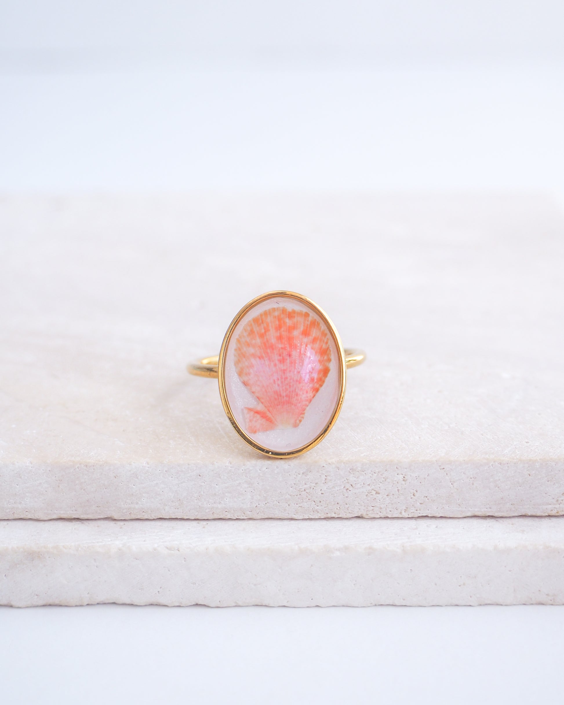 Ring with a pink and gold design on a white fabric background. Tiny Scallop Shell Gold Ring on stone displayed. Tiny Coral Pink Scallop Shell Bezel Ring, 18k gold stainless steel, handpicked scallop shell from the beach of Portugal.