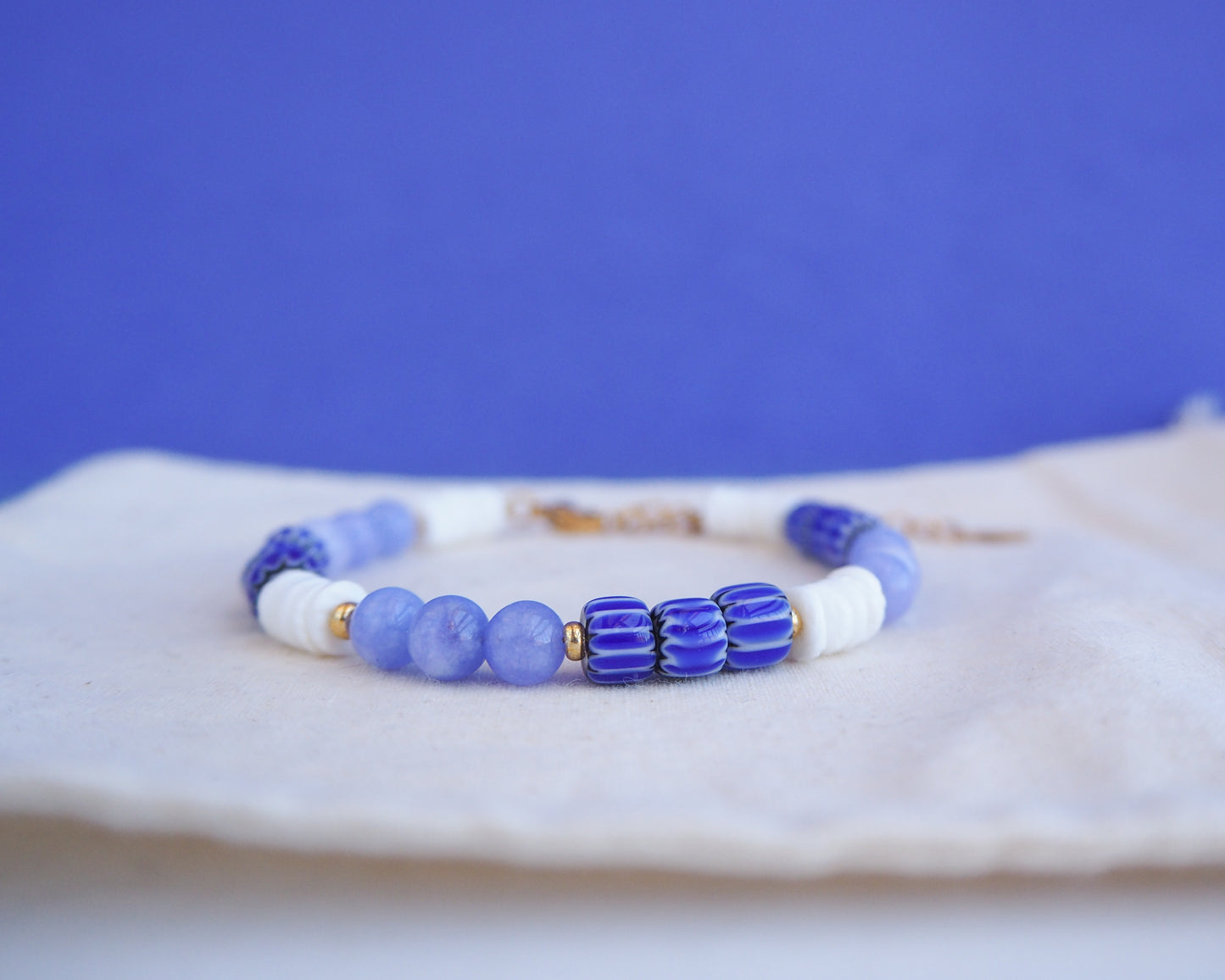 Close up of a beaded bracelet with blue and white beads on a blue background. Bracelet with  blue angelite gemstones, dark blue striped beads and white shell beads displayed on a light cotton and blue background.  