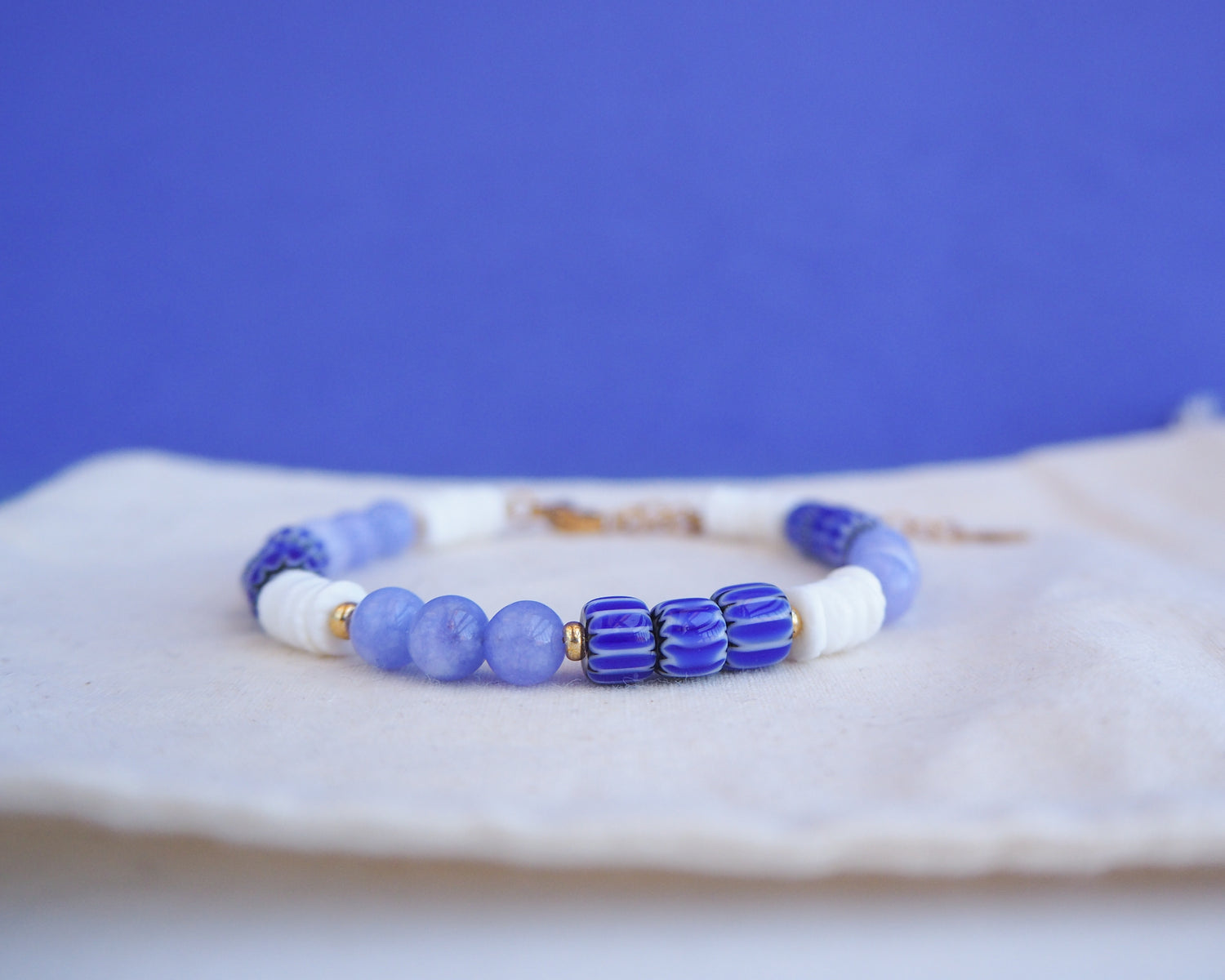 Close up of a beaded bracelet with blue and white beads on a blue background. Bracelet with  blue angelite gemstones, dark blue striped beads and white shell beads displayed on a light cotton and blue background.  