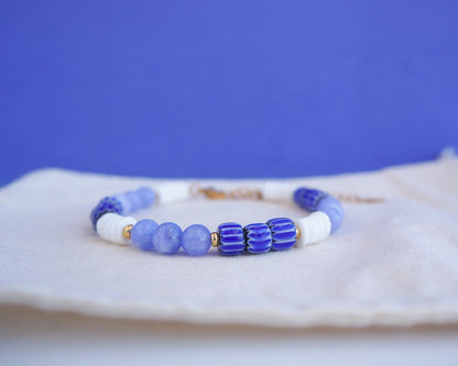 Close up of a beaded bracelet with blue and white beads on a blue background. Bracelet with  blue angelite gemstones, dark blue striped beads and white shell beads displayed on a light cotton and blue background.  