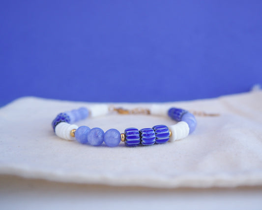 Close up of a beaded bracelet with blue and white beads on a blue background. Bracelet with  blue angelite gemstones, dark blue striped beads and white shell beads displayed on a light cotton and blue background.  