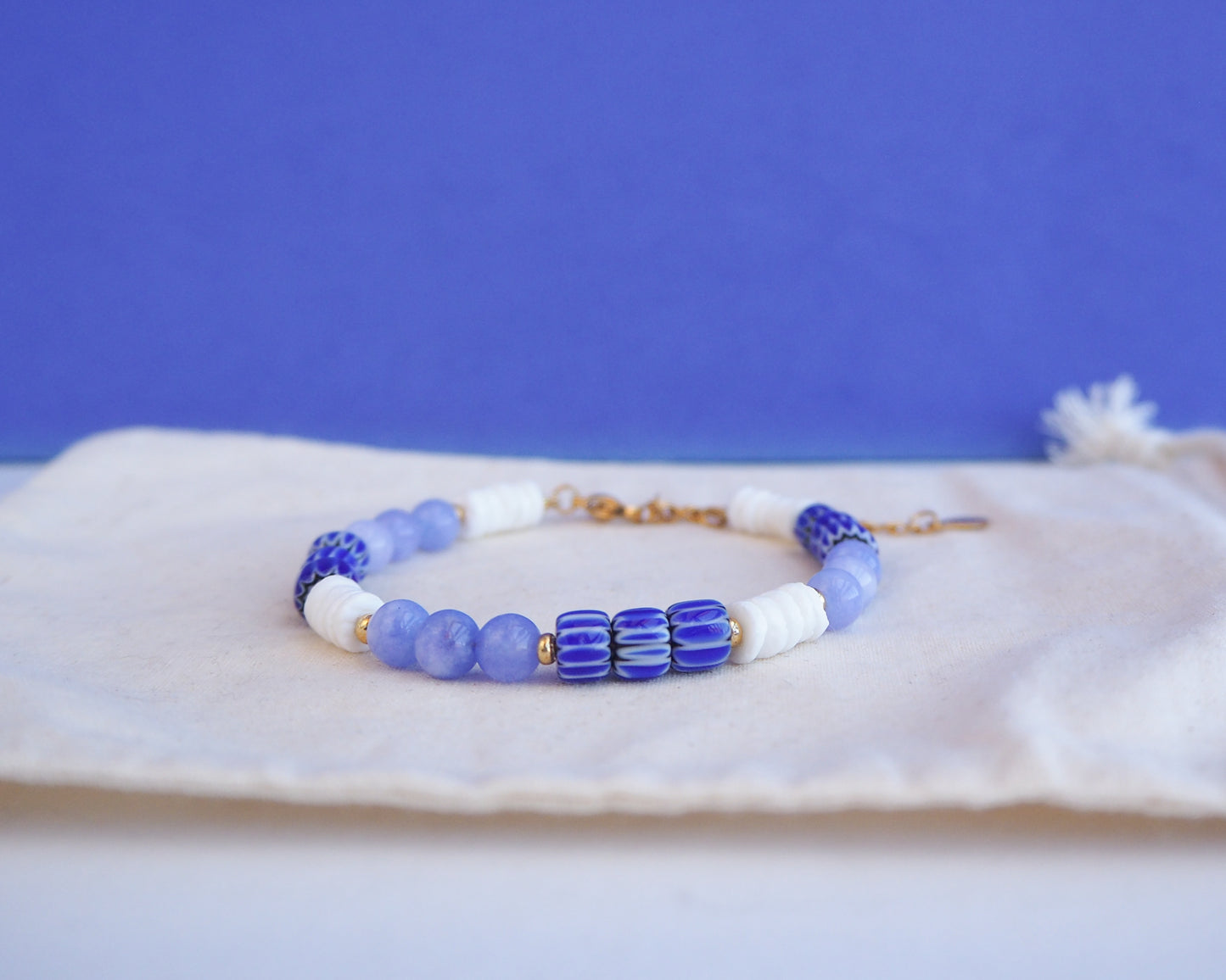 Beaded bracelet with blue and white beads on a blue background. Bracelet with  blue angelite gemstones, dark blue striped beads and white shell beads displayed on a light cotton and blue background.  