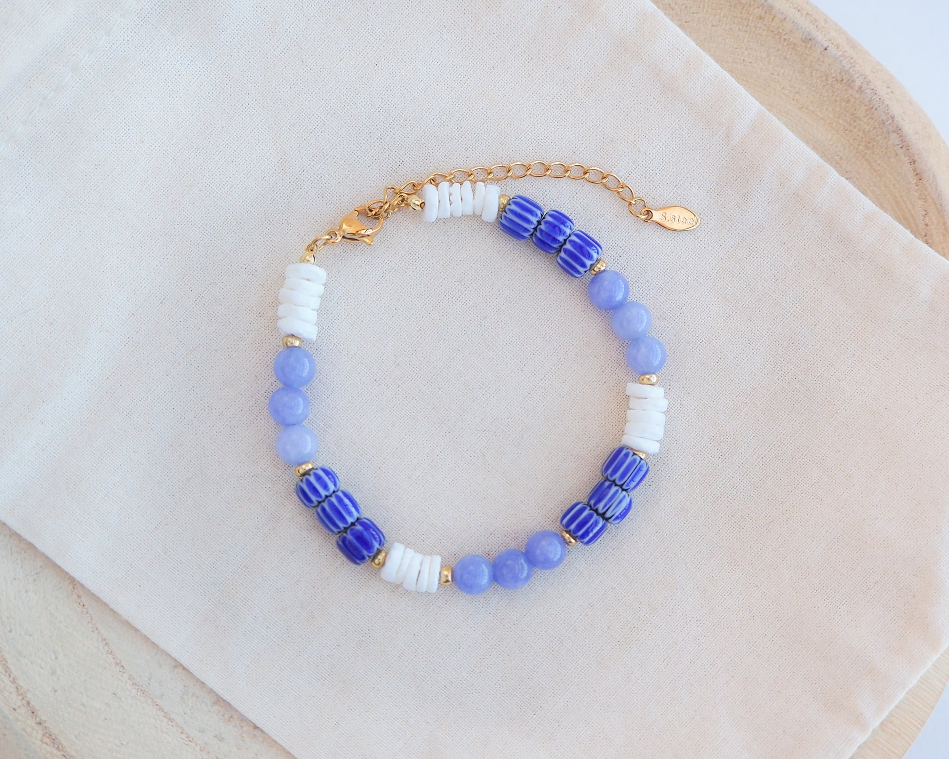 Beaded bracelet with blue, white, and gold beads on a beige fabric background. Front view of Bracelet with blue angelite gemstones, dark blue striped beads and white shell beads displayed on a light cotton .
