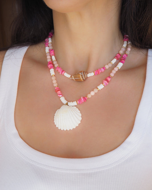 Necklace with pink and white beads and a shell pendant on a person wearing a white top. Model wearing a necklace with a big white cockle shell and a necklace with a big cone shell paired with rose quartz gemstone beads, pink stones and white shell beads. The shell is a mediterranean cockle shell and Mediterranean Cone Shell from Portugal and is about 4 cm. The color of the shell is ivory and brown.