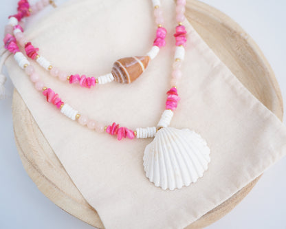 Side view of a necklace with pink beads and a white shell pendant on a light background. Necklace with pink and white beads and a shell pendant on light cotton displayed. A big white cockle shell and a necklace with a big cone shell paired with rose quartz gemstone beads, pink stones and white shell beads. The shell is a mediterranean cockle shell and Mediterranean Cone Shell from Portugal and is about 4 cm. The color of the shell is ivory and brown.