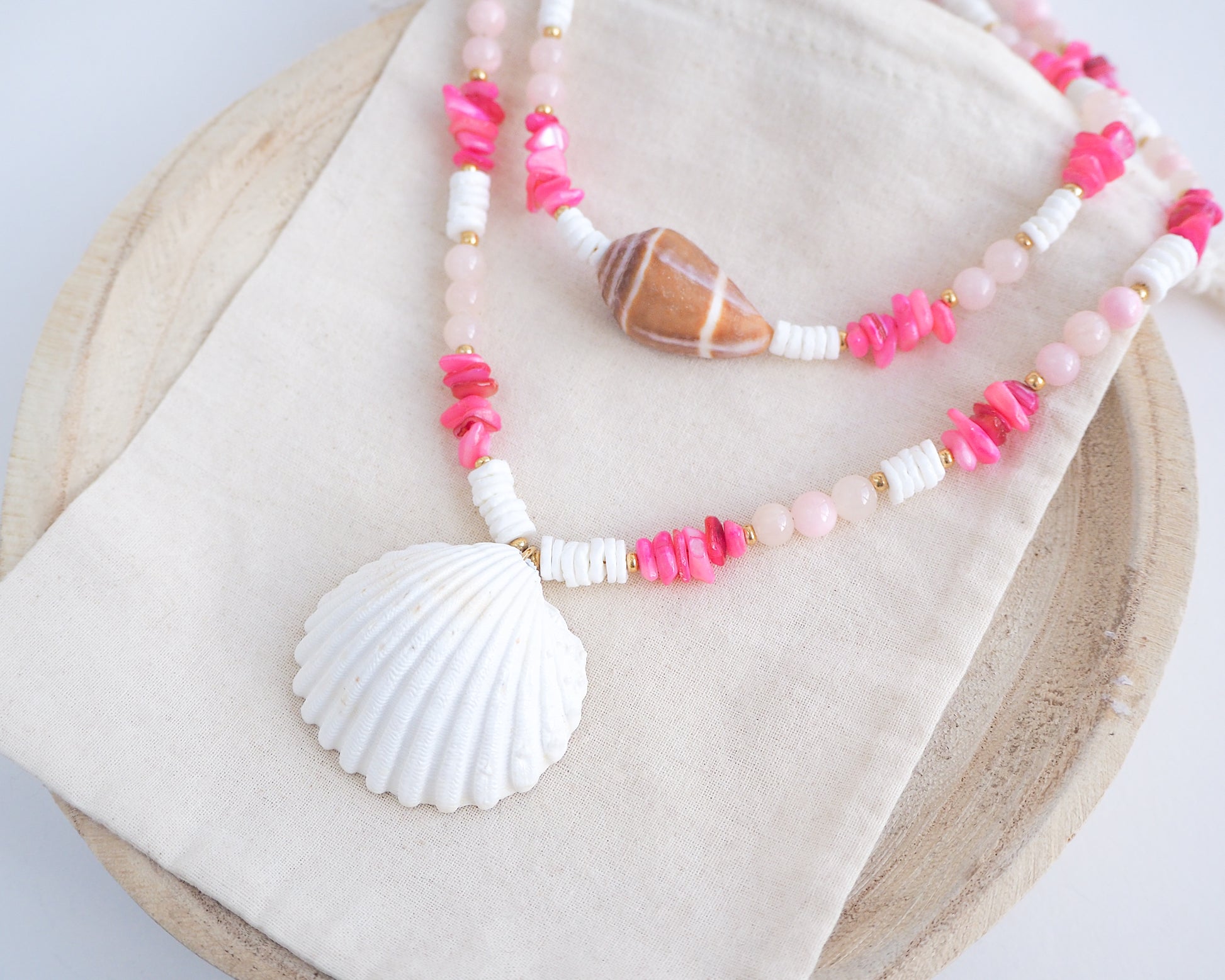 Necklace with pink and white beads and a shell pendant on light cotton displayed. A big white cockle shell and a necklace with a big cone shell paired with rose quartz gemstone beads, pink stones and white shell beads. The shell is a mediterranean cockle shell and Mediterranean Cone Shell from Portugal and is about 4 cm. The color of the shell is ivory and brown.