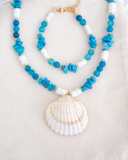 Necklace with blue and white beads and a shell pendant on a light background. Front view of A necklace with a big clam shell paired with agate gemstone beads, turquoise stones and white shell beads on cotton displayed. The shell is a mediterranean cockle shell from Portugal and is about 4 cm. The color of the shell is ivory bege. There is also a matching bracelet on the cotton dipslayed