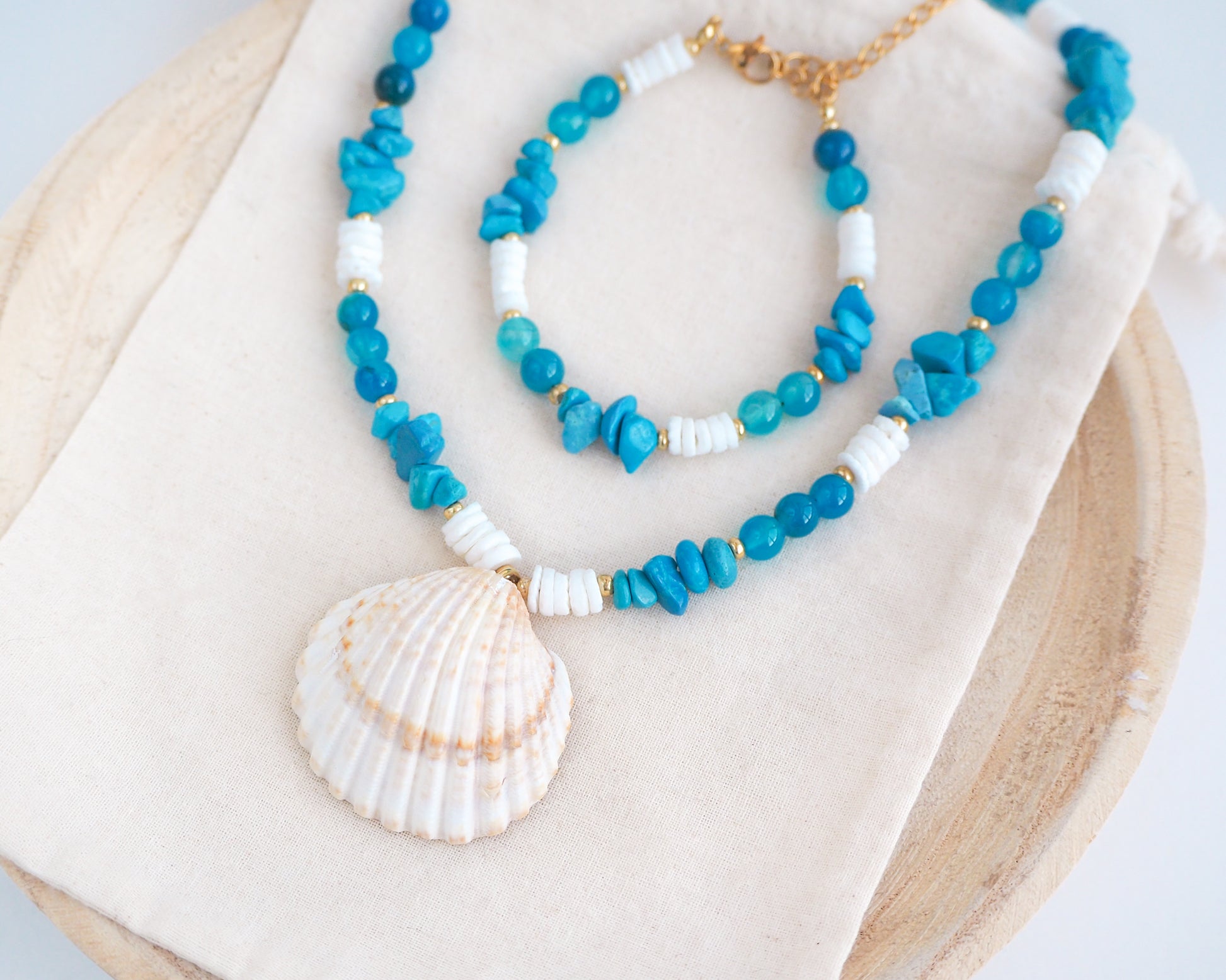 Side view of a necklace with a big clam shell paired with agate gemstone beads, turquoise stones and white shell beads on cotton displayed. The shell is a mediterranean cockle shell from Portugal and is about 4 cm. The color of the shell is ivory bege. There is also a matching bracelet on the cotton dipslayed