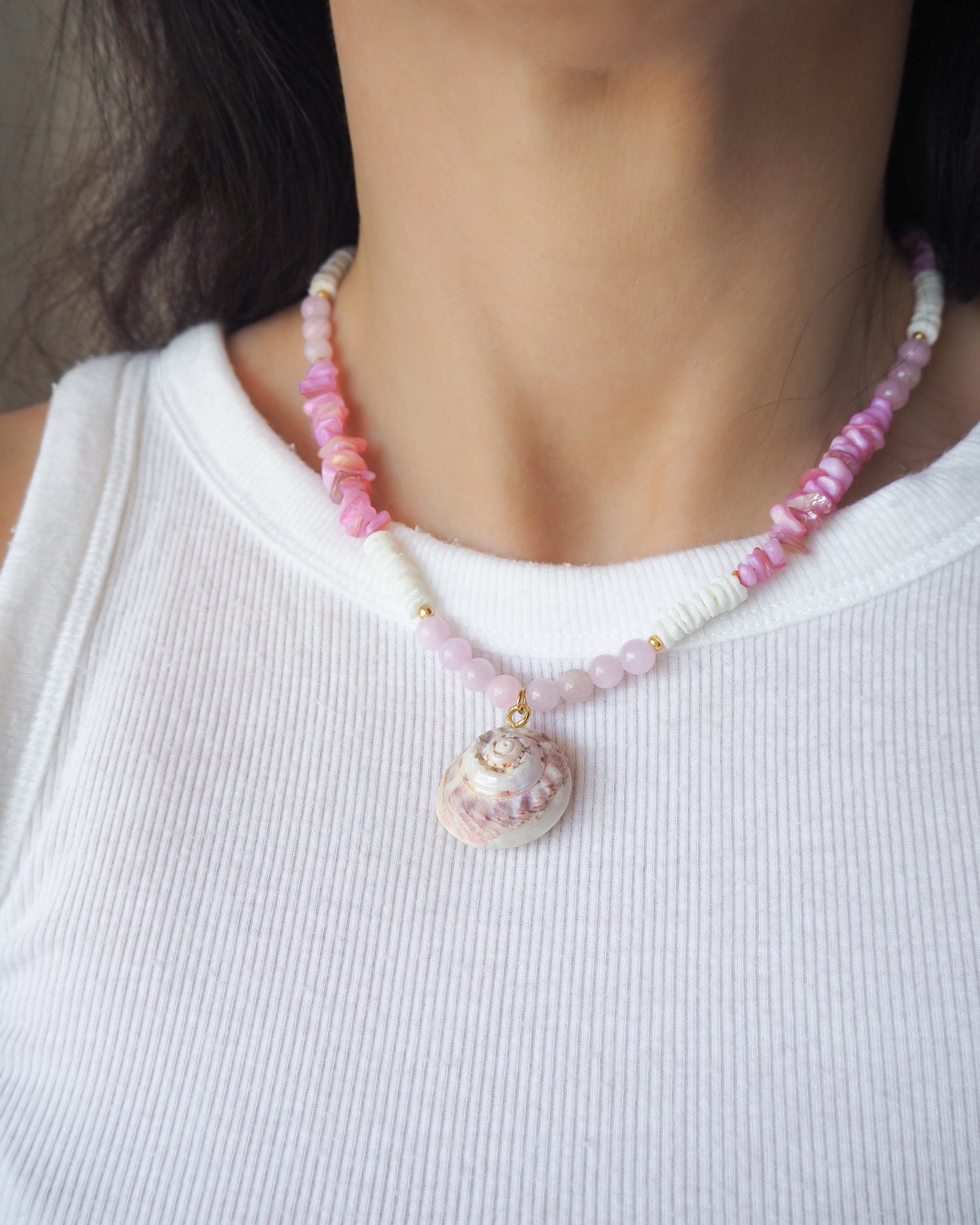 Person wearing a pink shell beaded necklace with a shell pendant on a white background.  A necklace with a big pink magus top shell paired with rose quartz gemstone beads, soft pink stones and white shell beads. The shell is a magus top shell handpicked from Portugal and is about 3 cm. The color of the shell is soft pink.