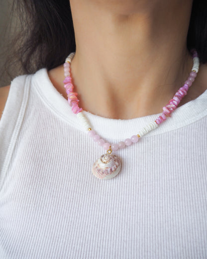 Person wearing a pink shell beaded necklace with a shell pendant on a white background.  A necklace with a big pink magus top shell paired with rose quartz gemstone beads, soft pink stones and white shell beads. The shell is a magus top shell handpicked from Portugal and is about 3 cm. The color of the shell is soft pink.