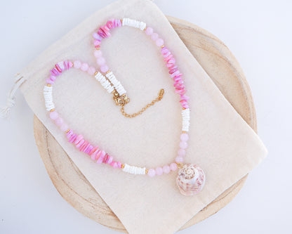 Beaded necklace with pink and white beads on a beige fabric background. A necklace with a big pink magus top shell paired with rose quartz gemstone beads, soft pink stones and white shell beads. The shell is a magus top shell handpicked from Portugal and is about 3 cm. The color of the shell is soft pink.