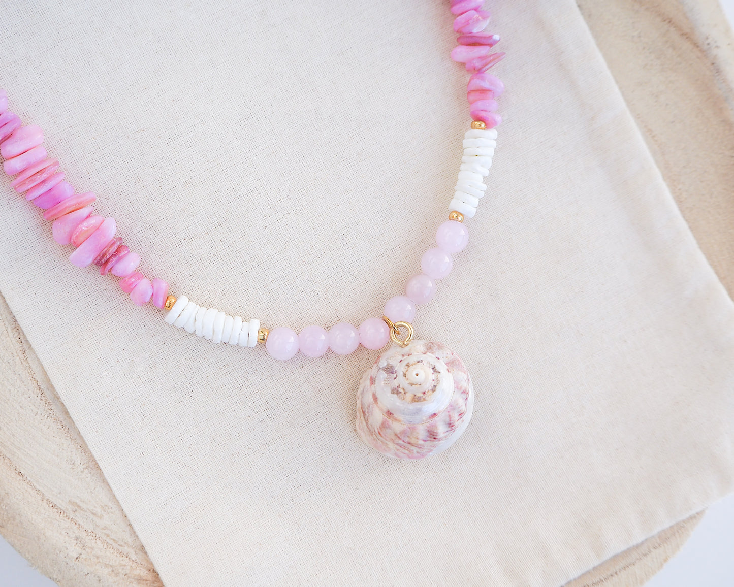 Necklace with pink and white beads and a shell pendant on a light background. A necklace with a big pink magus top shell paired with rose quartz gemstone beads, soft pink stones and white shell beads. The shell is a magus top shell handpicked from Portugal and is about 3 cm. The color of the shell is soft pink.