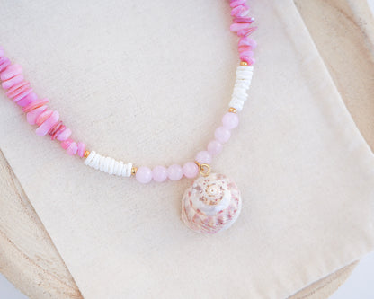 Necklace with pink and white beads and a shell pendant on a light background. A necklace with a big pink magus top shell paired with rose quartz gemstone beads, soft pink stones and white shell beads. The shell is a magus top shell handpicked from Portugal and is about 3 cm. The color of the shell is soft pink.
