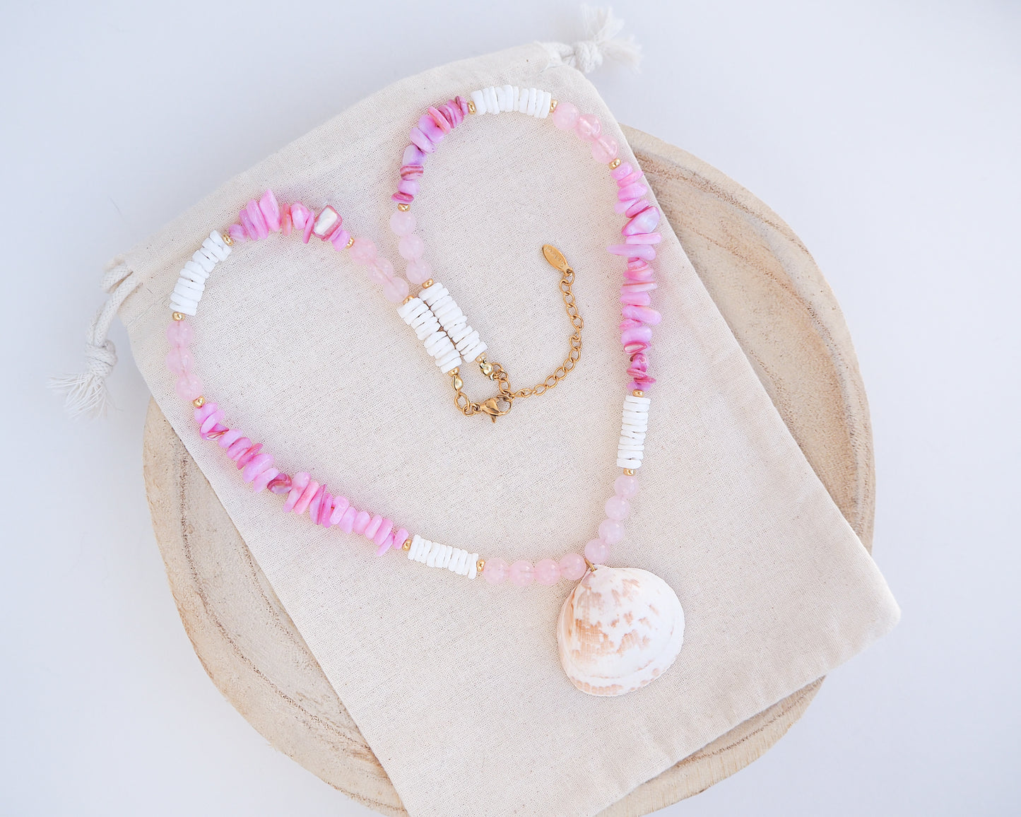 A necklace with pink and white beads and a shell pendant on light cotton displayed. A necklace with a big white cockle shell paired with rose quartz gemstone beads, soft pink stones and white shell beads. The shell is a Norwegian cockle shell handpicked from Portugal and is about 4 cm. The color of the shell is ivory white with a beige brown pattern.