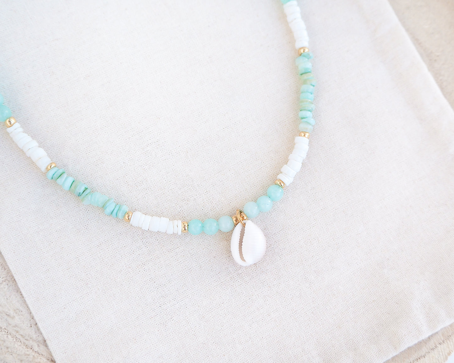 Necklace with turquoise and white beads on a light background. Close up of a cowrie necklace with soft green stone beads, jade beads and white shell beads. The center piece of this necklace is a Northern Cowrie Shell, handpicked from the beach of Portugal. The shell is white and about 1 cm. 