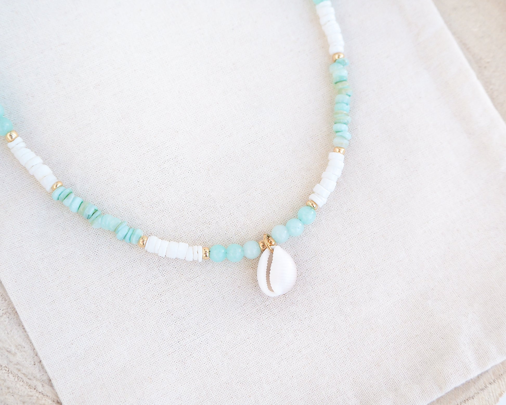 Necklace with turquoise and white beads on a light background. Close up of a cowrie necklace with soft green stone beads, jade beads and white shell beads. The center piece of this necklace is a Northern Cowrie Shell, handpicked from the beach of Portugal. The shell is white and about 1 cm. 