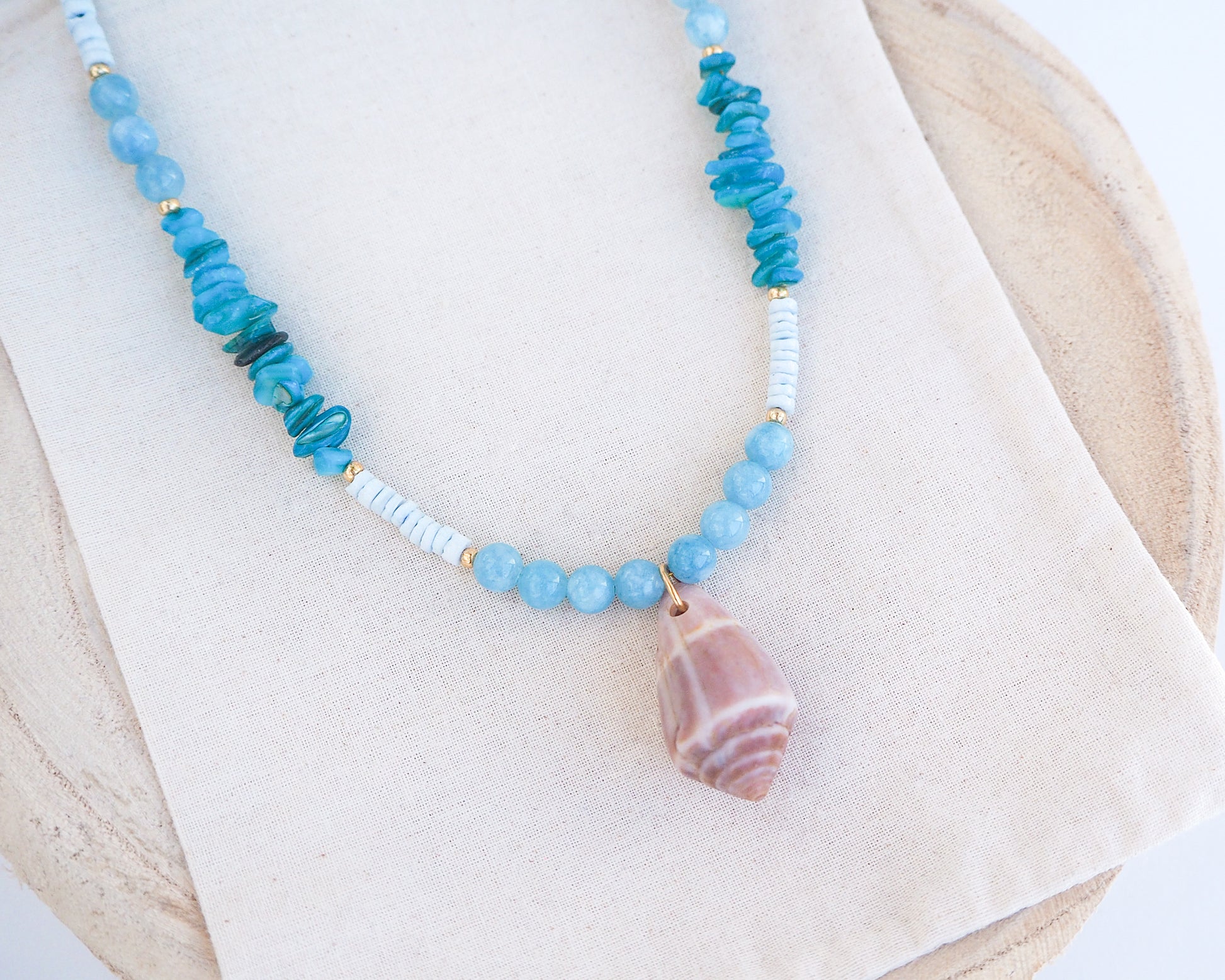 Close up of necklace with blue and white beads and a large cone shell pendant on a light background. A necklace with a mediterranean cone shell paired with aquamarine gemstone beads and white shell beads. The shell is a mediterranean cone shell from Portugal and is about 4 cm. The color of the shell is brown.