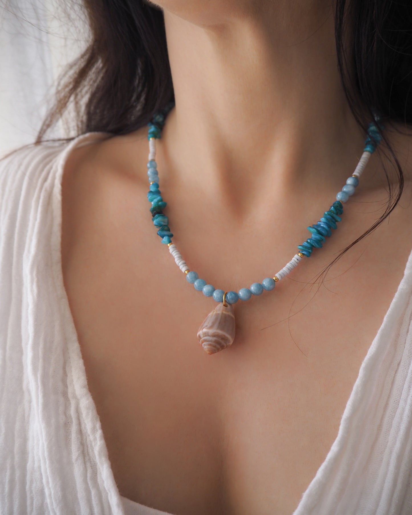 Model wearing a necklace with a big cone shell paired with aquamarine gemstone beads, turquoise stones and light blue shell beads. The shell is a mediterranean cone shell from Portugal and is about 4 cm. The color of the shell is brown.