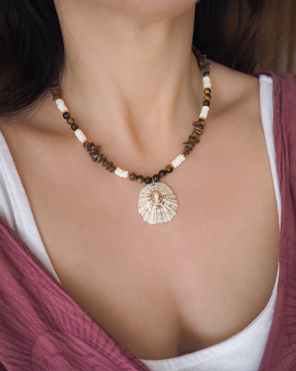 Necklace with shell pendant worn by a person. Big limpet necklace with brown and white beads on display. Shell is paired with tiger eye gemstone beads, brown stones and white shell beads. The shell is a rough limpet shell handpicked from Portugal and is about 4 cm. The color of the shell is ivory white with a beige brown pattern.