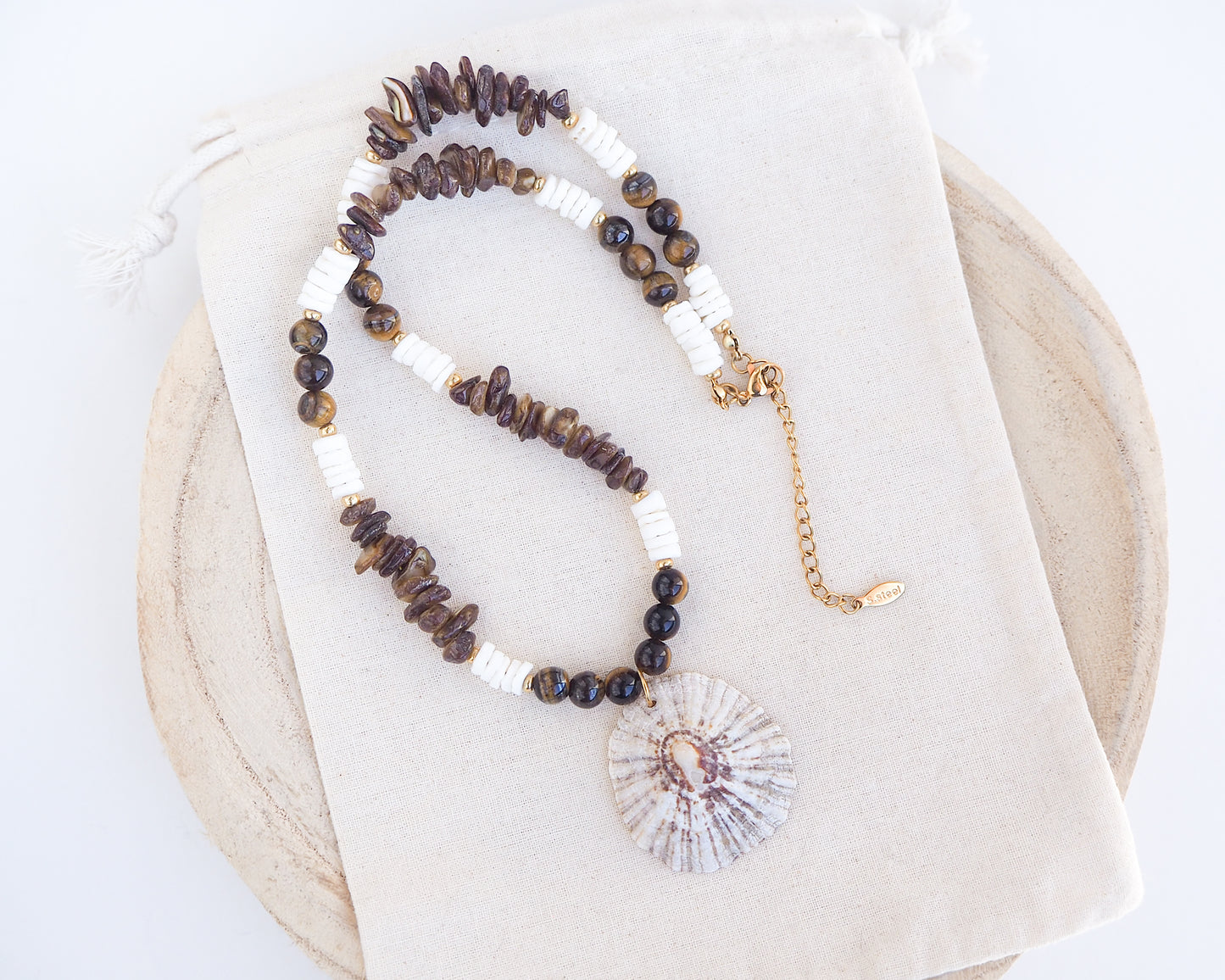 Beaded necklace with a shell pendant on a white background. Big limpet necklace with brown and white beads on display. Shell is paired with tiger eye gemstone beads, brown stones and white shell beads. The shell is a rough limpet shell handpicked from Portugal and is about 4 cm. The color of the shell is ivory white with a beige brown pattern.