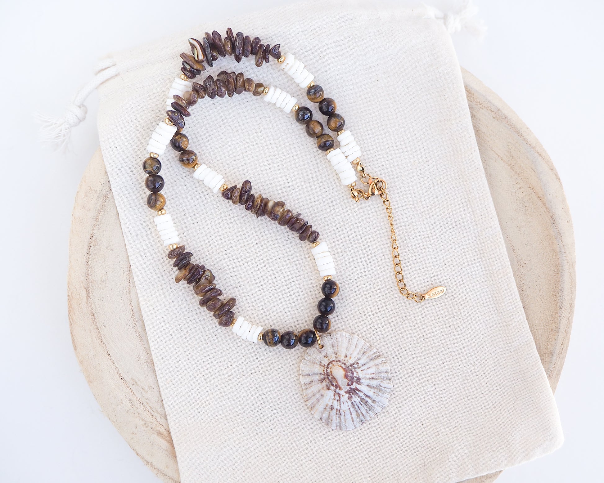Beaded necklace with a shell pendant on a white background. Big limpet necklace with brown and white beads on display. Shell is paired with tiger eye gemstone beads, brown stones and white shell beads. The shell is a rough limpet shell handpicked from Portugal and is about 4 cm. The color of the shell is ivory white with a beige brown pattern.