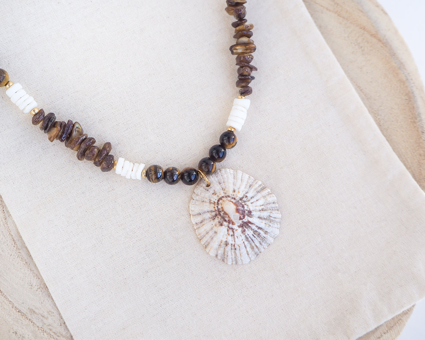 Big limpet necklace with brown and white beads on display. Shell is paired with tiger eye gemstone beads, brown stones and white shell beads. The shell is a rough limpet shell handpicked from Portugal and is about 4 cm. The color of the shell is ivory white with a beige brown pattern.