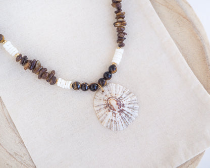 Big limpet necklace with brown and white beads on display. Shell is paired with tiger eye gemstone beads, brown stones and white shell beads. The shell is a rough limpet shell handpicked from Portugal and is about 4 cm. The color of the shell is ivory white with a beige brown pattern.