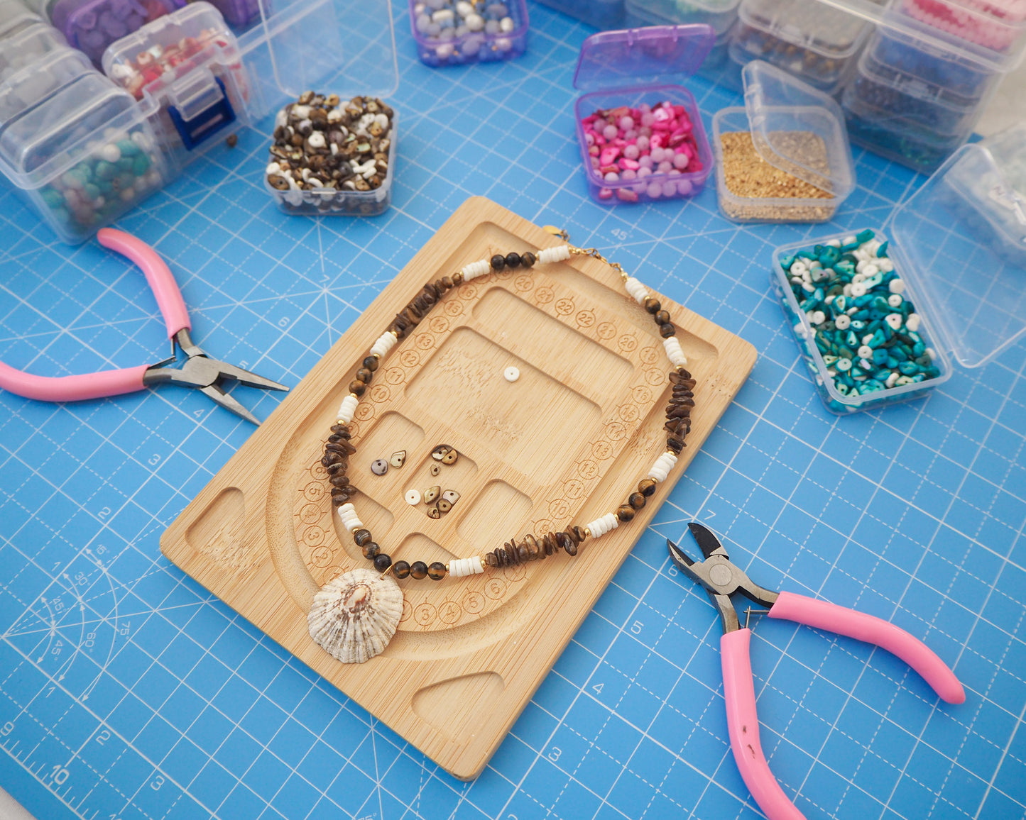 Jewelry-making setup with beads, tools, and a necklace on a blue cutting mat. Making of a big limpet necklace with brown and white beads in process. Shell is paired with tiger eye gemstone beads, brown stones and white shell beads. The shell is a rough limpet shell handpicked from Portugal and is about 4 cm. The color of the shell is ivory white with a beige brown striped pattern. 