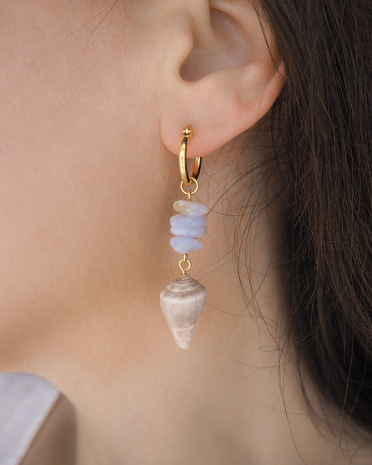 Gold hoop earring with shell and stone pendant worn by a person. Gold Cone Shell Blue Gemstone Drop Earrings in ear model. Gold earring hoops with real Mediterranean Cone Shells from Portugal and blue agate gemstones. Made with 14k gold stainless steel hoop earrings.   