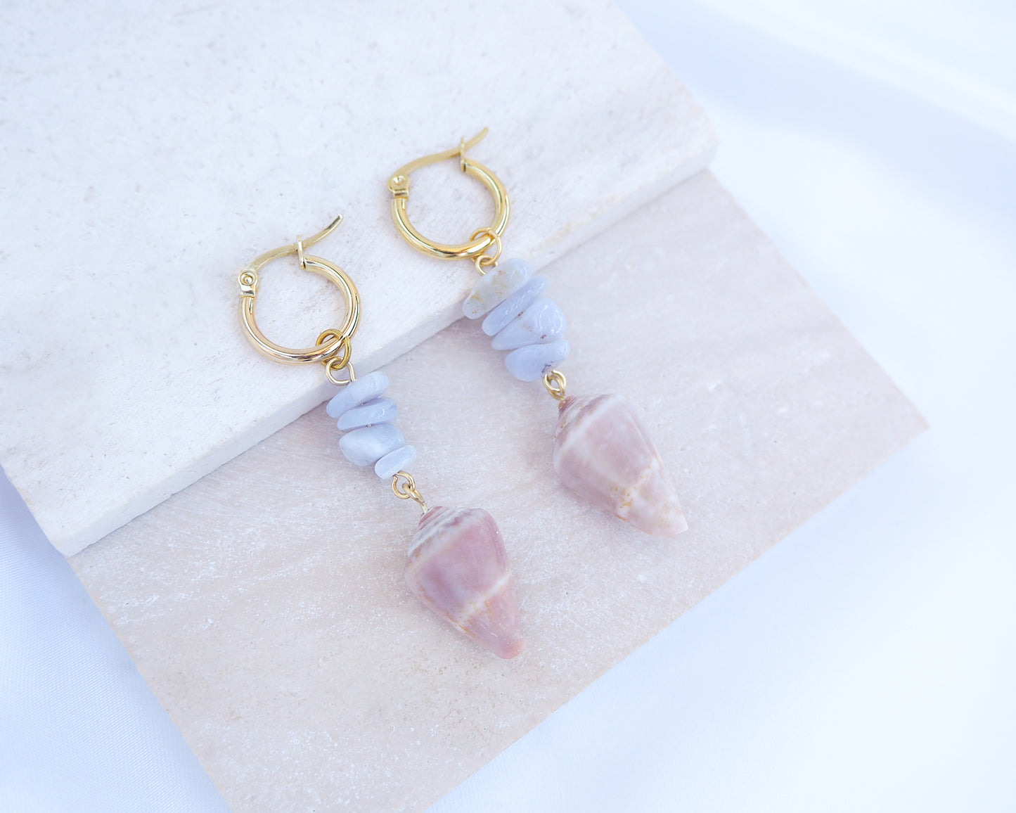 Gold Cone Shell Blue Gemstone Earrings displayed on light stone. Gold earring hoops with real Mediterranean Cone Shells from Portugal and blue agate gemstones. Made with 14k gold stainless steel hoop earrings.   