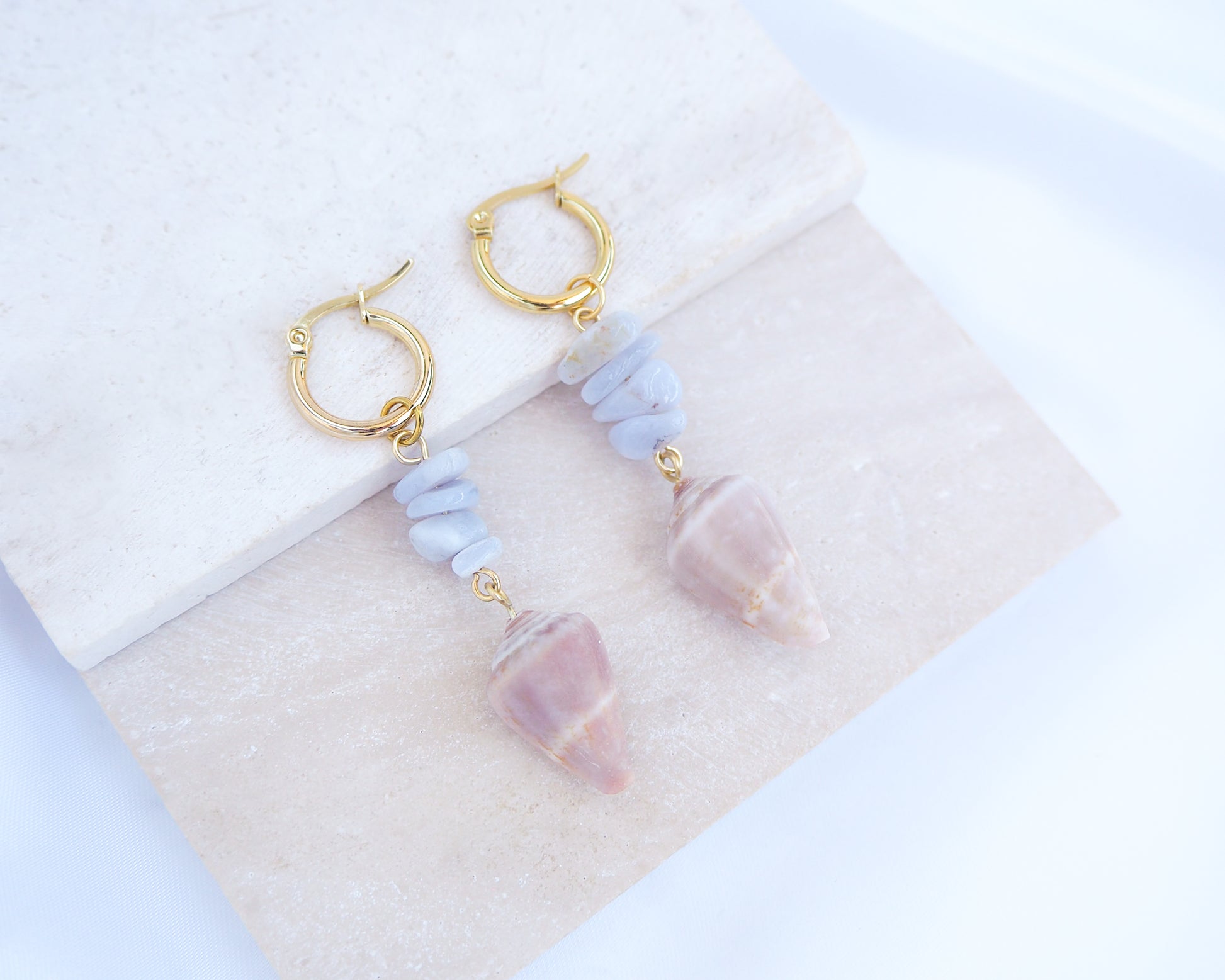 Gold Cone Shell Blue Gemstone Earrings displayed on light stone. Gold earring hoops with real Mediterranean Cone Shells from Portugal and blue agate gemstones. Made with 14k gold stainless steel hoop earrings.   