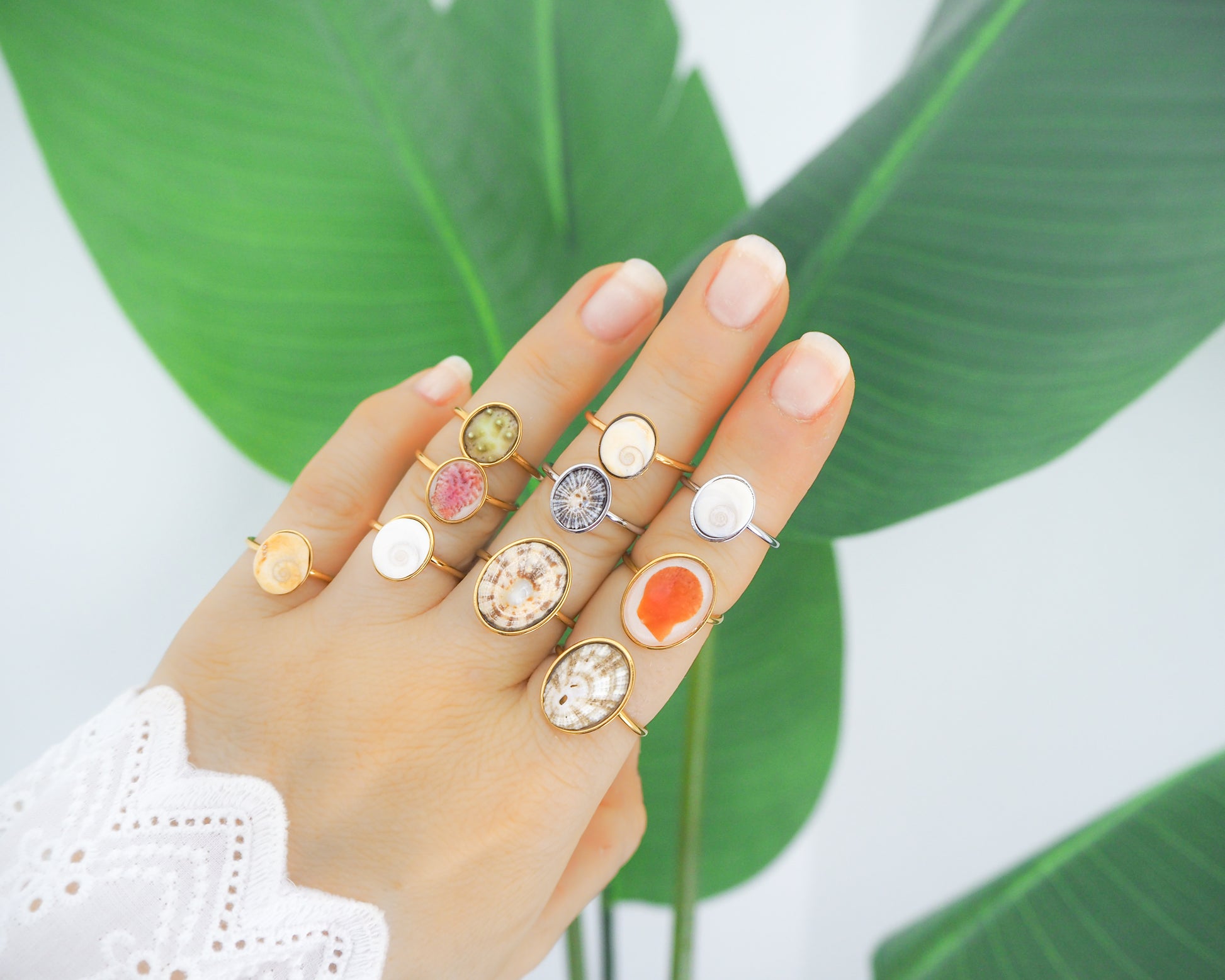 Hand wearing multiple rings with colorful gemstones against a green leaf background. All made with handpicked shells from Portugal set in a bezel. Adjustable silver or gold stainless steel rings.