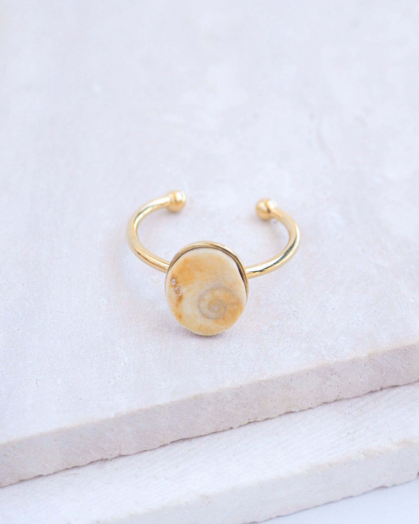 Side view Gold bezel ring with a tiny beige shiva eye on stone displayed with light background. Tiny beige Eye of Saint Lucia Gold Ring on stone displayed, 18k gold stainless steel shiva eye operculum, Saint Lucy Eye, operculum pendant