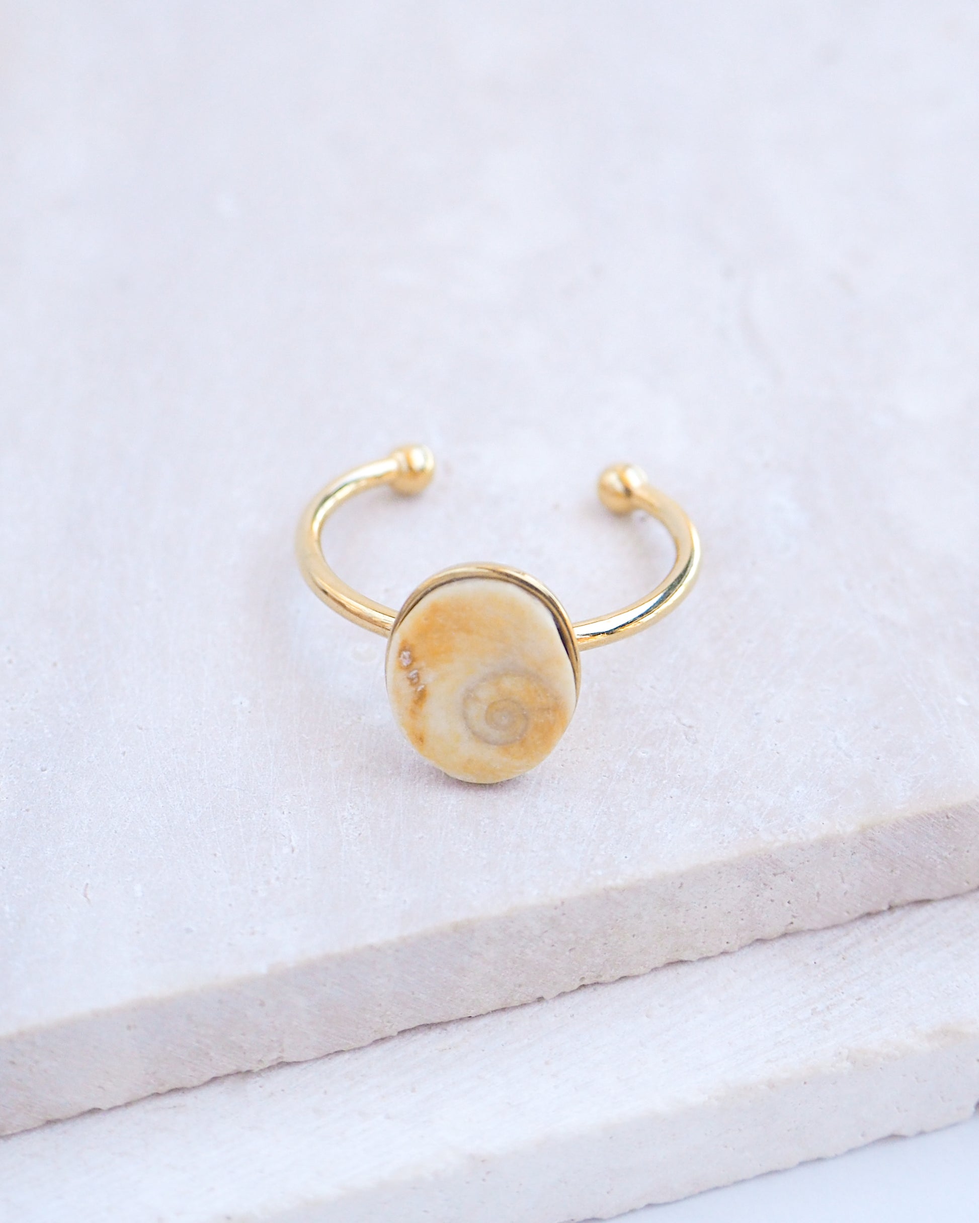 Side view Gold bezel ring with a tiny beige shiva eye on stone displayed with light background. Tiny beige Eye of Saint Lucia Gold Ring on stone displayed, 18k gold stainless steel shiva eye operculum, Saint Lucy Eye, operculum pendant