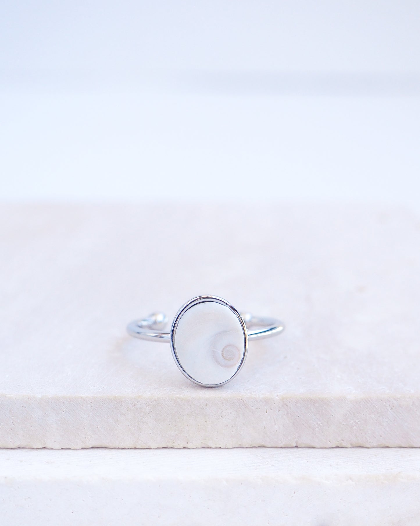 Silver bezel ring with a tiny ivory white shiva eye on stone displayed with light background. White Eye of Saint Lucia Gold Ring on stone displayed, silver stainless steel shiva eye operculum, Saint Lucy Eye, operculum pendant