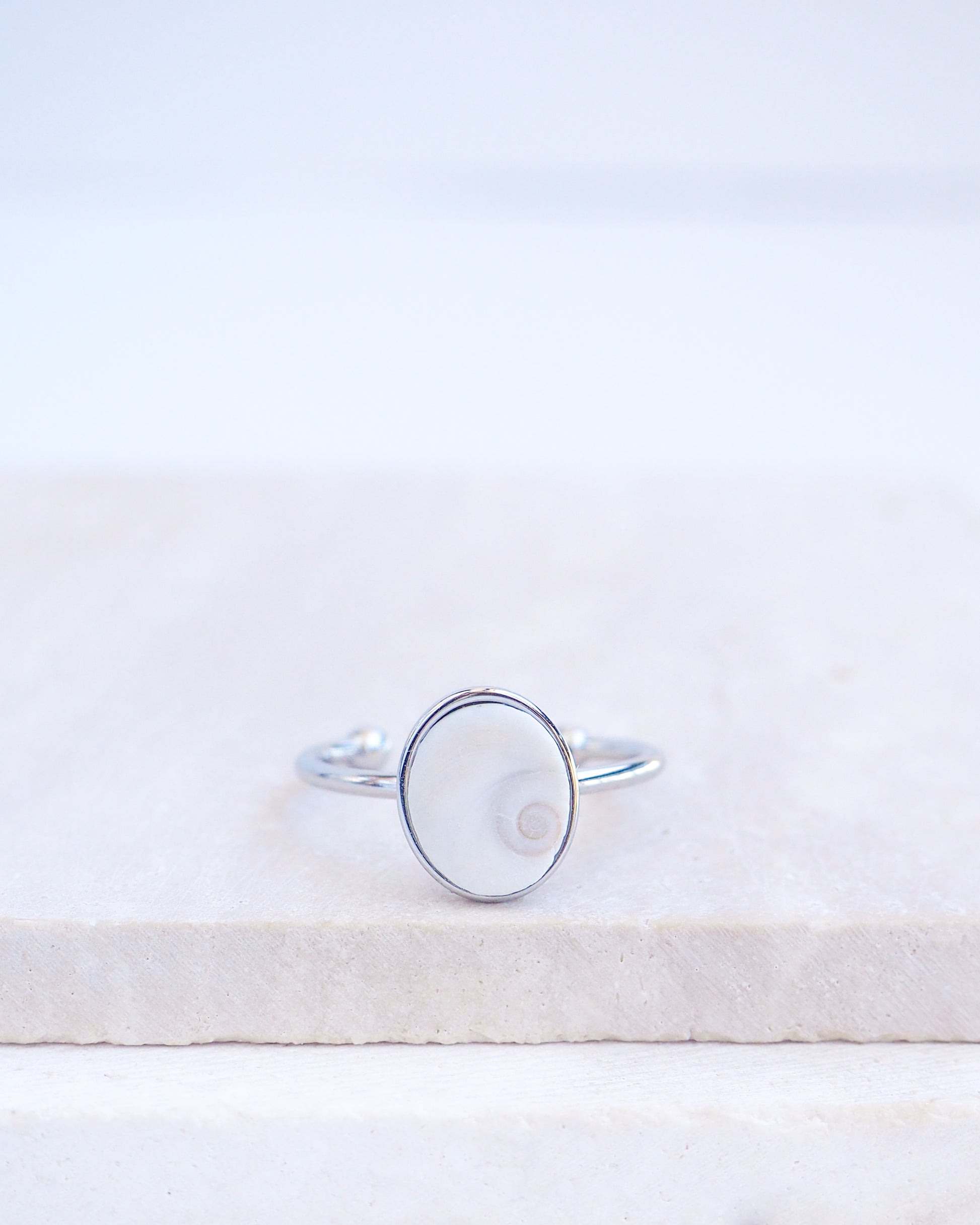 Silver bezel ring with a tiny ivory white shiva eye on stone displayed with light background. White Eye of Saint Lucia Gold Ring on stone displayed, silver stainless steel shiva eye operculum, Saint Lucy Eye, operculum pendant