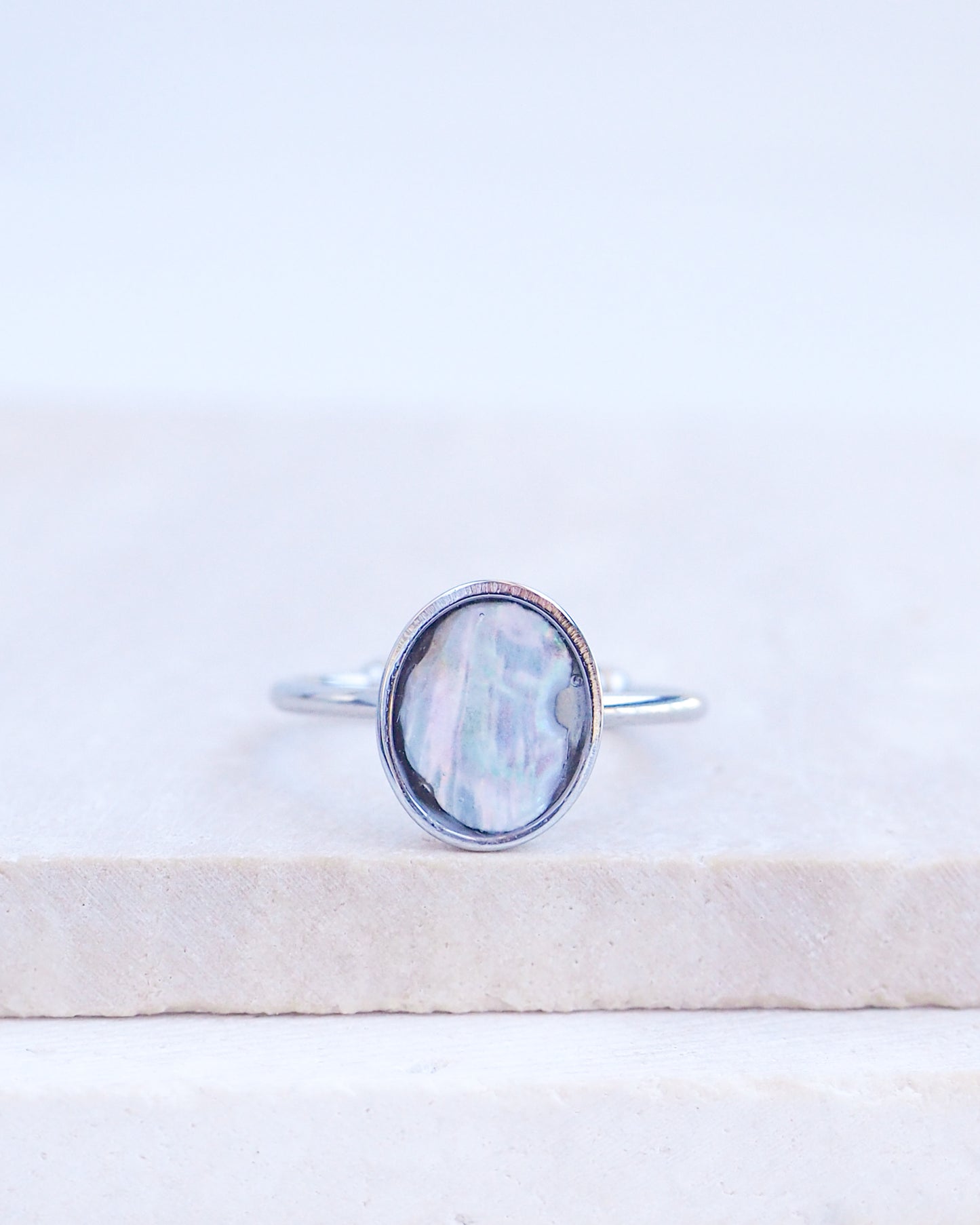 A silver ring with an oval iridescent mother-of-pearl piece of abalone shell, resting on a light beige stone slab against a white background. Silver ring with a piece of abalone shell. The piece of abalone shell was found on the beaches of Portugal. Set in a bezel ring made of silver stainless steel. The ring is adjustable. 