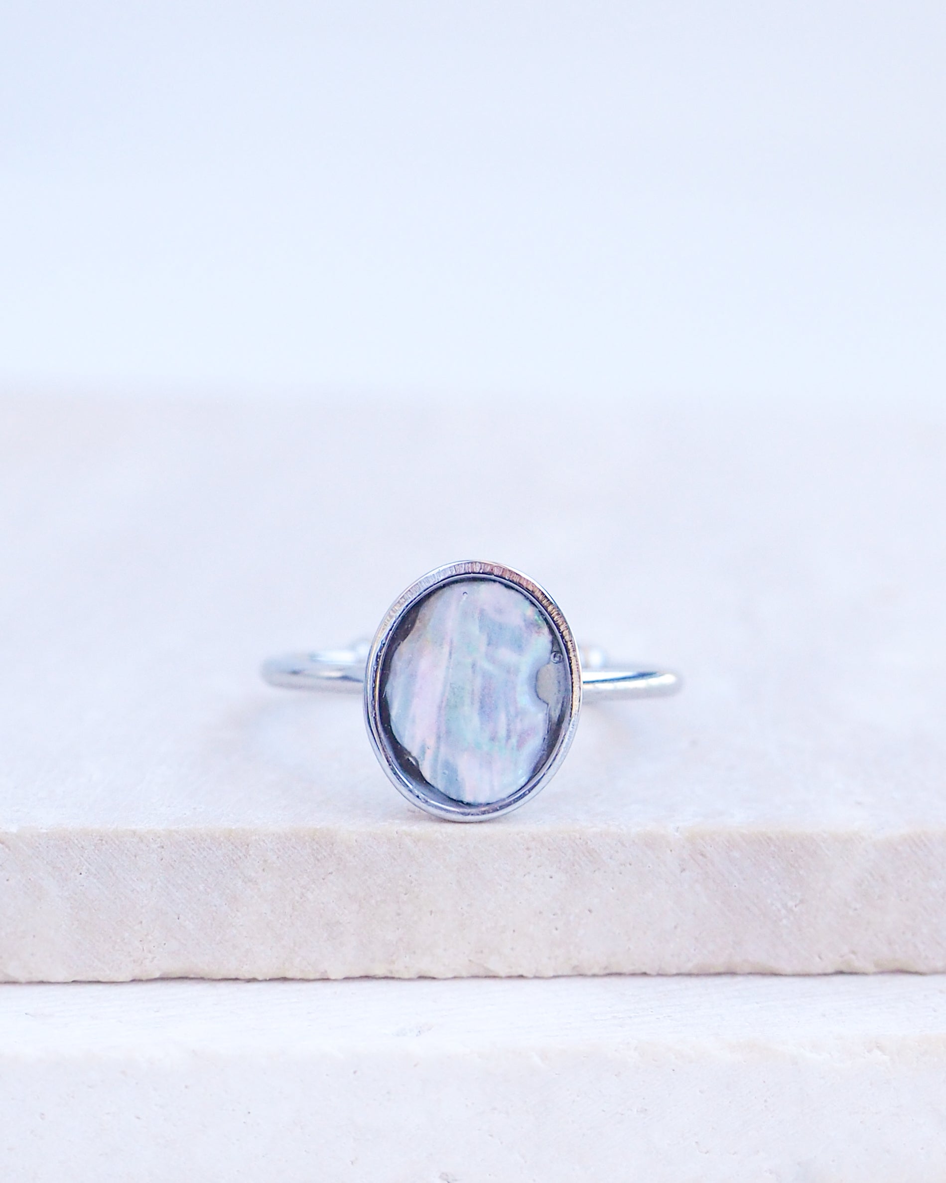 A silver ring with an oval iridescent mother-of-pearl piece of abalone shell, resting on a light beige stone slab against a white background. Silver ring with a piece of abalone shell. The piece of abalone shell was found on the beaches of Portugal. Set in a bezel ring made of silver stainless steel. The ring is adjustable. 