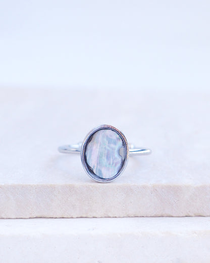 A silver ring with an oval iridescent mother-of-pearl piece of abalone shell, resting on a light beige stone slab against a white background. Silver ring with a piece of abalone shell. The piece of abalone shell was found on the beaches of Portugal. Set in a bezel ring made of silver stainless steel. The ring is adjustable. 