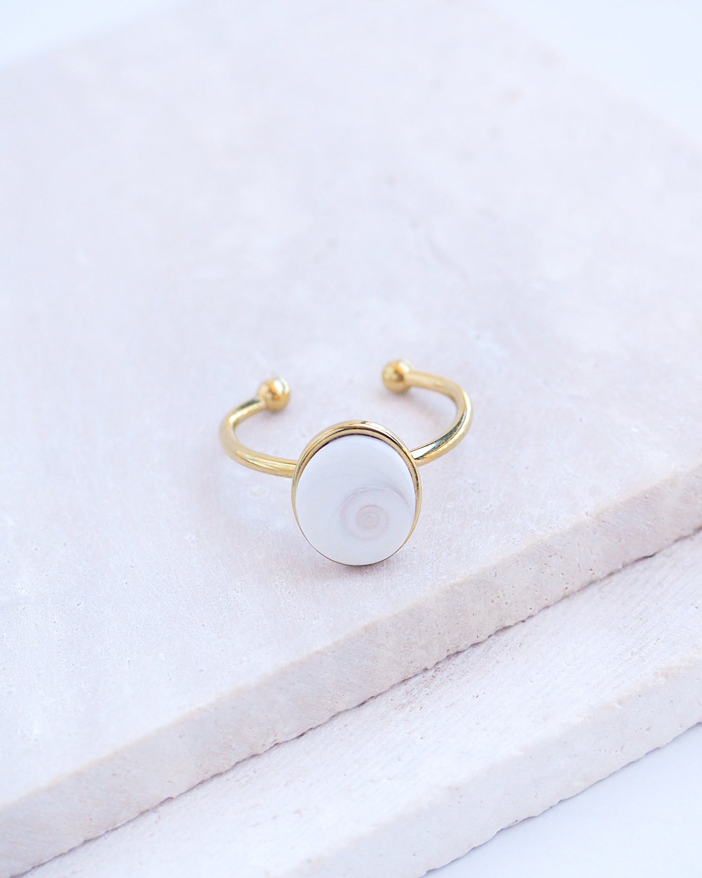 Side view of Gold bezel ring with a white shiva eye on stone displayed with light background. White Eye of Saint Lucia Gold Ring on stone displayed, 18k gold stainless steel shiva eye operculum, Saint Lucy Eye, operculum pendant