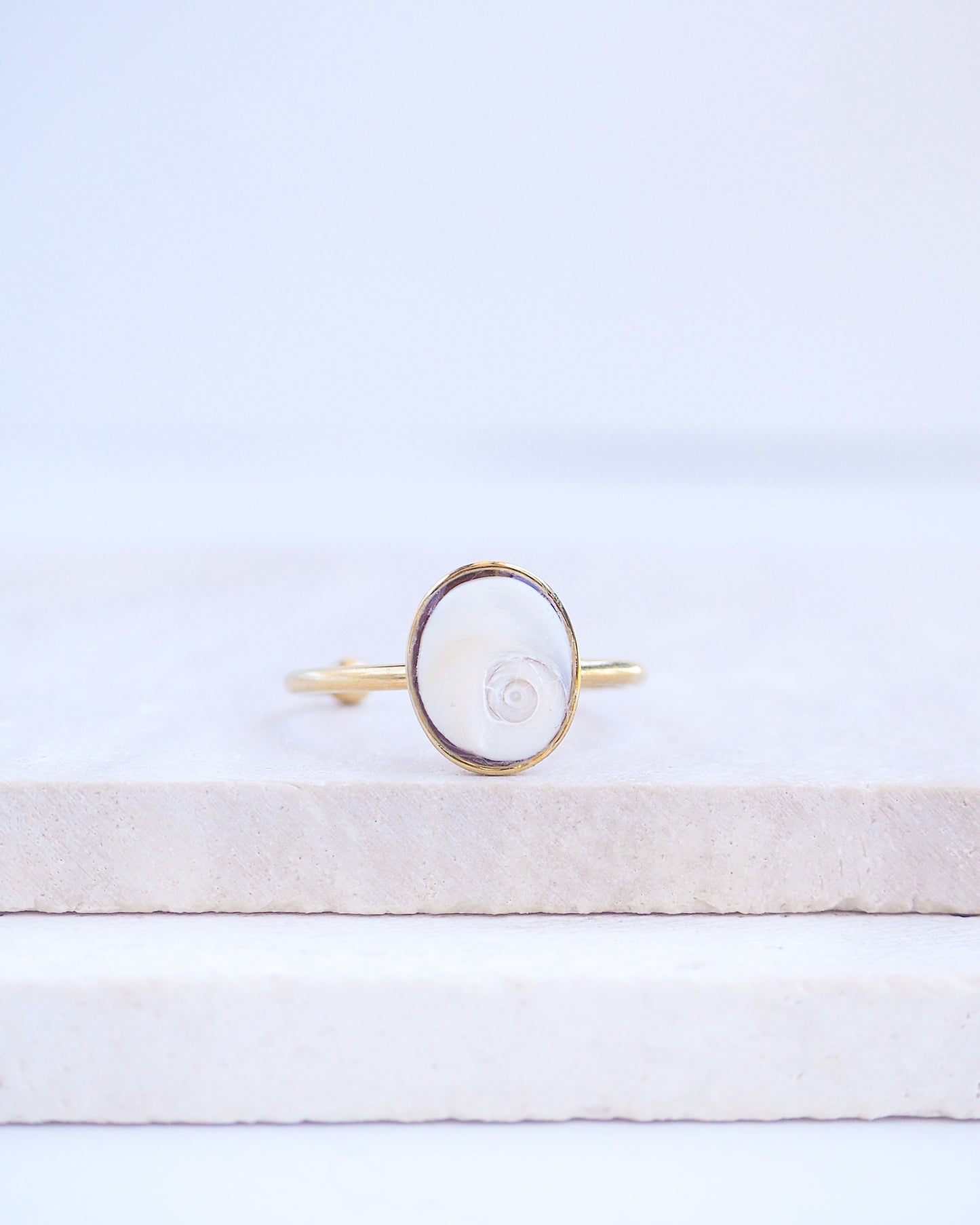 Front view of Gold bezel ring with a tiny ivory white shiva eye on stone displayed with light background. White Eye of Saint Lucia Gold Ring on stone displayed, 18k gold stainless steel shiva eye operculum, Saint Lucy Eye, operculum pendant