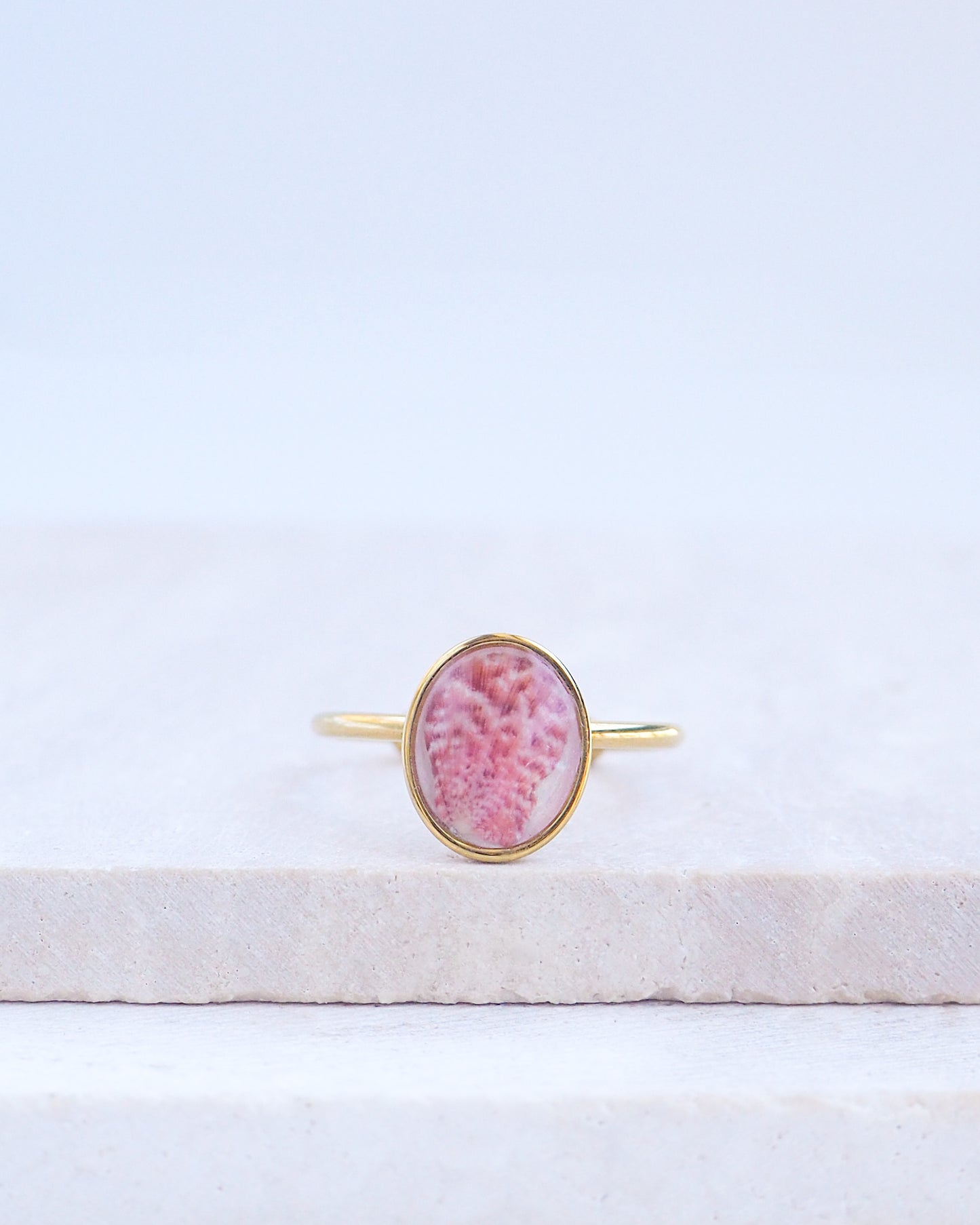 Ring with a pink and gold design on a white fabric background. Tiny Scallop Shell Gold Ring on stone displayed. Tiny Pink Scallop Shell Bezel Ring, 18k gold stainless steel, handpicked scallop shell from the beach of Portugal.