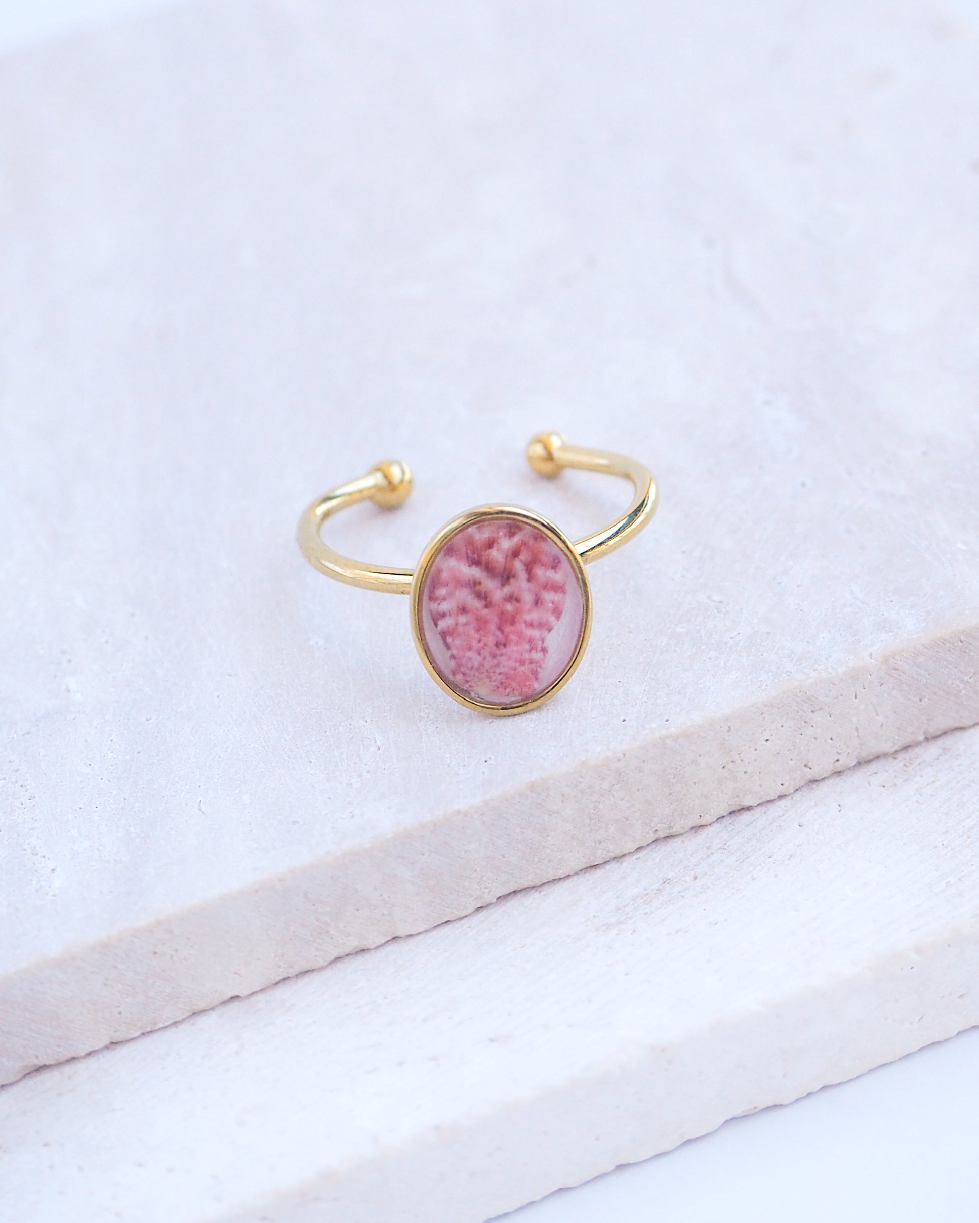 Ring with a pink and gold design on a white fabric background. Tiny Scallop Shell Gold Ring on stone displayed. Tiny Pink Scallop Shell Bezel Ring, 18k gold stainless steel, handpicked scallop shell from the beach of Portugal.