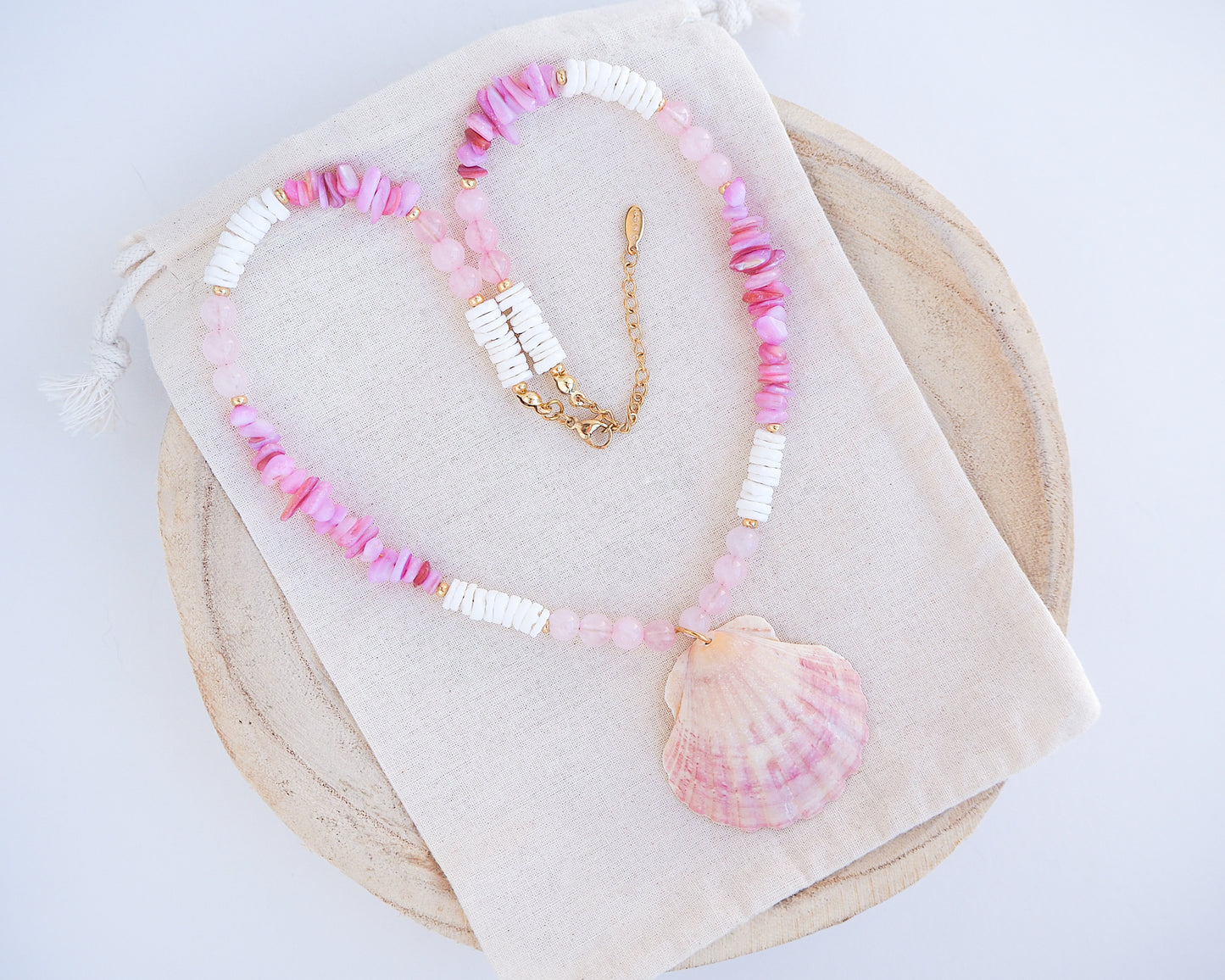 A necklace with pink and white beads and a shell pendant on light cotton displayed. A necklace with a big pink scallop shell paired with rose quartz gemstone beads, soft pink stones and white shell beads. The shell is a pilgrim's scallop shell handpicked from Portugal and is about 5 cm. The color of the shell is soft pink.